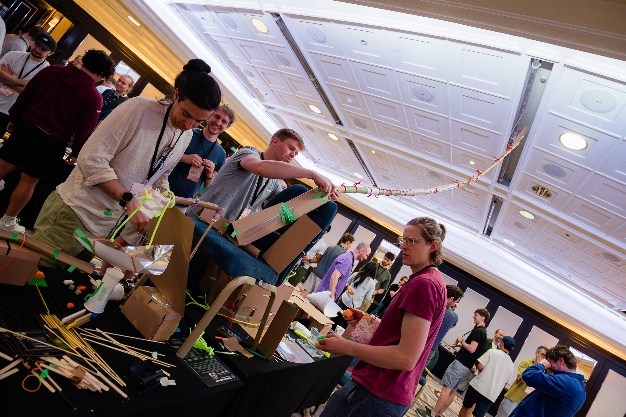 Long shot of a completed marble run mid-build · a team suspending the track from a chair back across the ballroom.