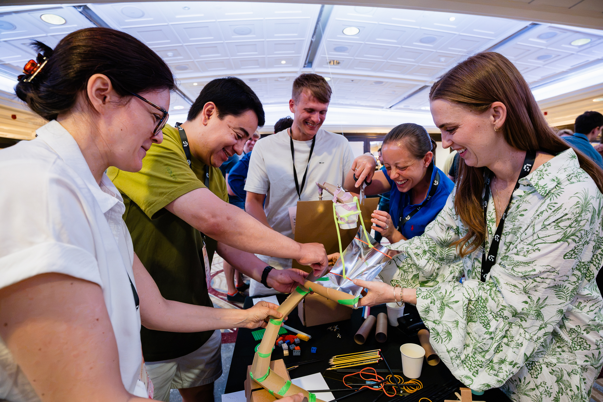 Iso Live finale · a team laughing as they cut and tape a foil pouch onto a cardboard marble run tower.