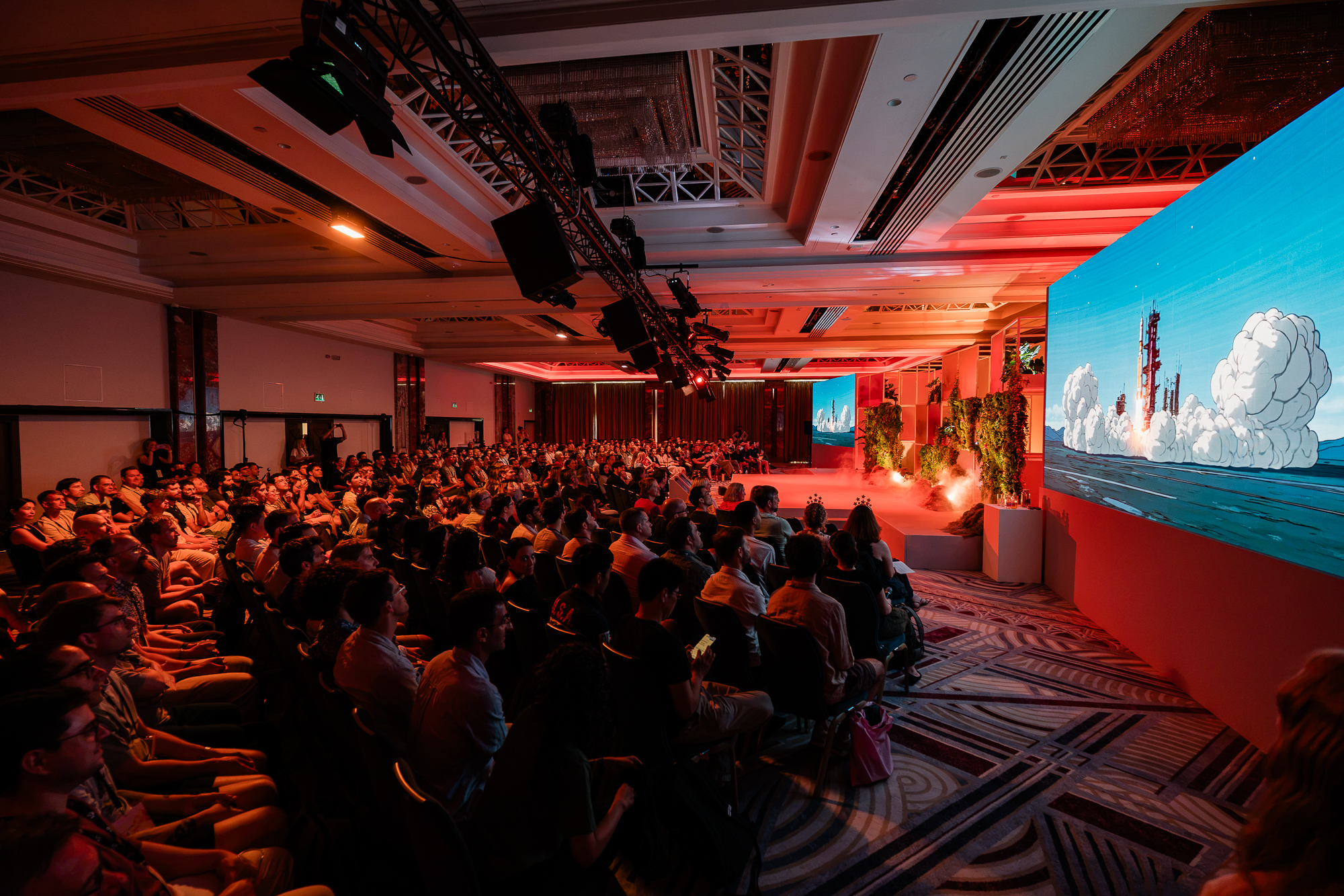 Iso Orbit reveal · packed ballroom watching the launch sequence on the widescreen with dramatic red uplight.