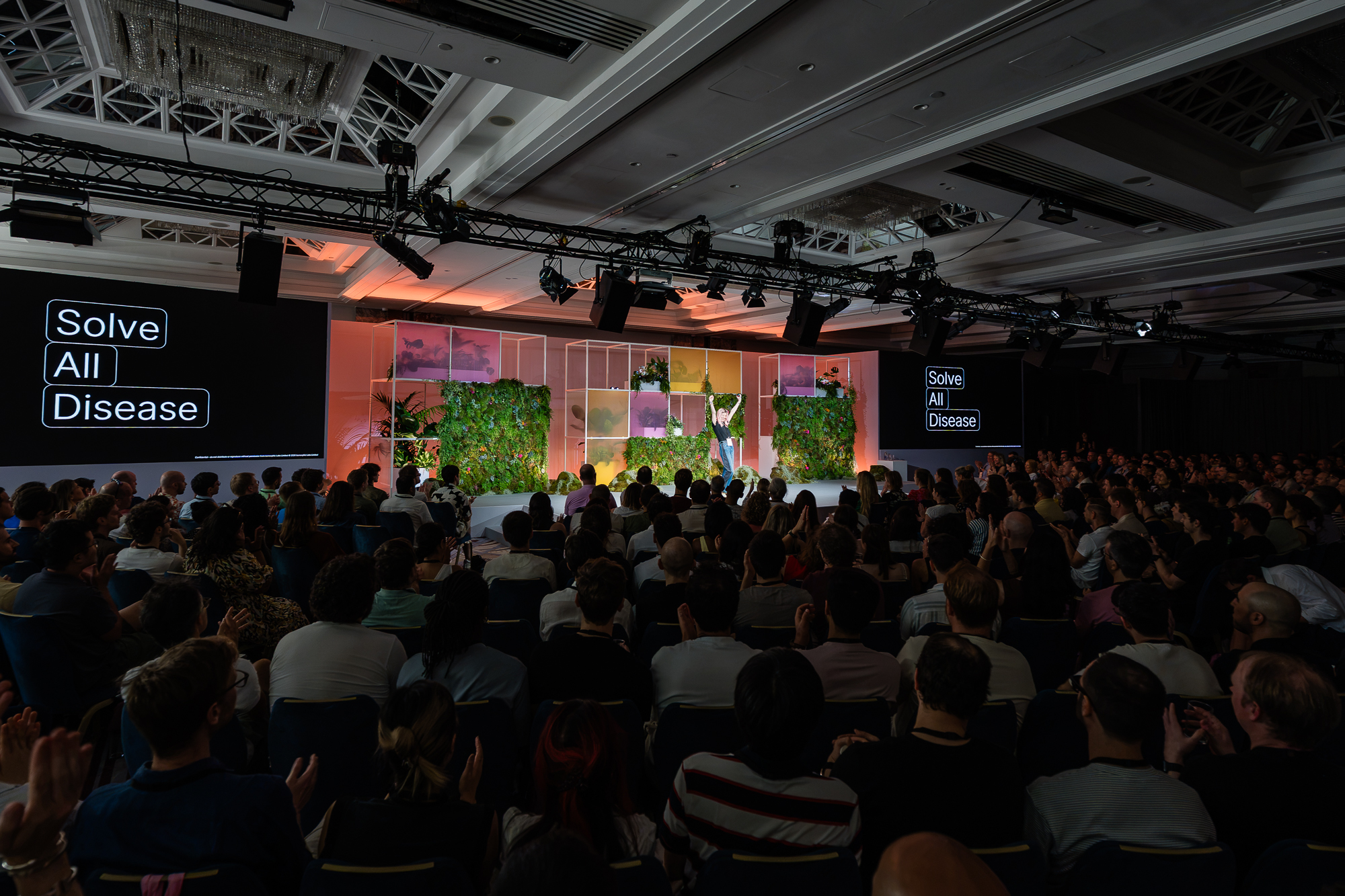 Iso Live 2026 main stage · Solve All Disease keynote on a steel grid and living foliage set, 400 colleagues in the audience.